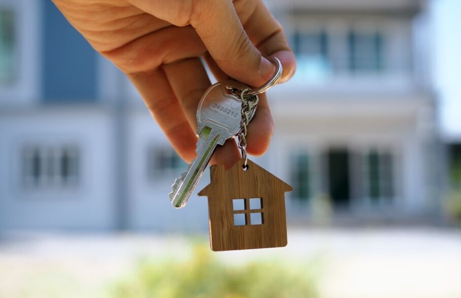 A hand holding a key with a house shaped keyring in front of a property